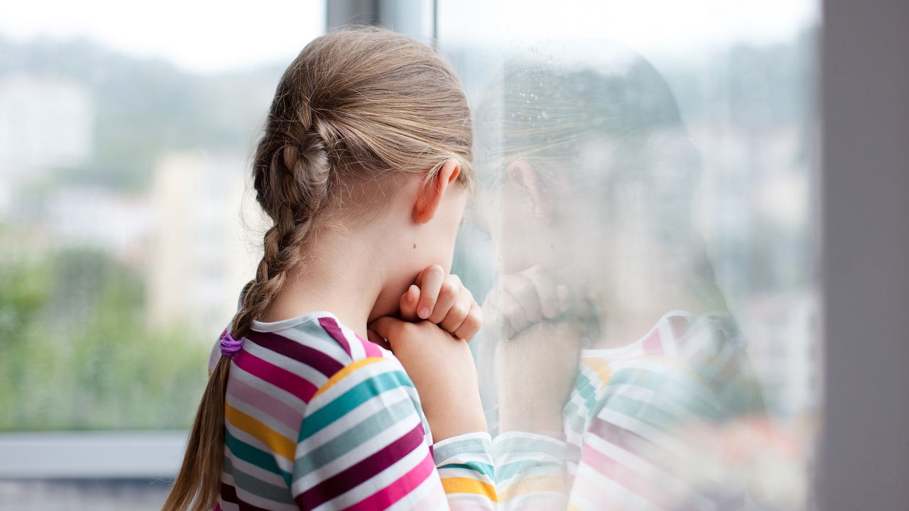 Sad child looking through rainy window at home. Upset kid in self isolation during quarantine.