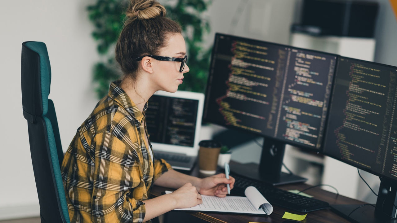 Confident female programmer working on software code using multiple screens in a modern office workspace