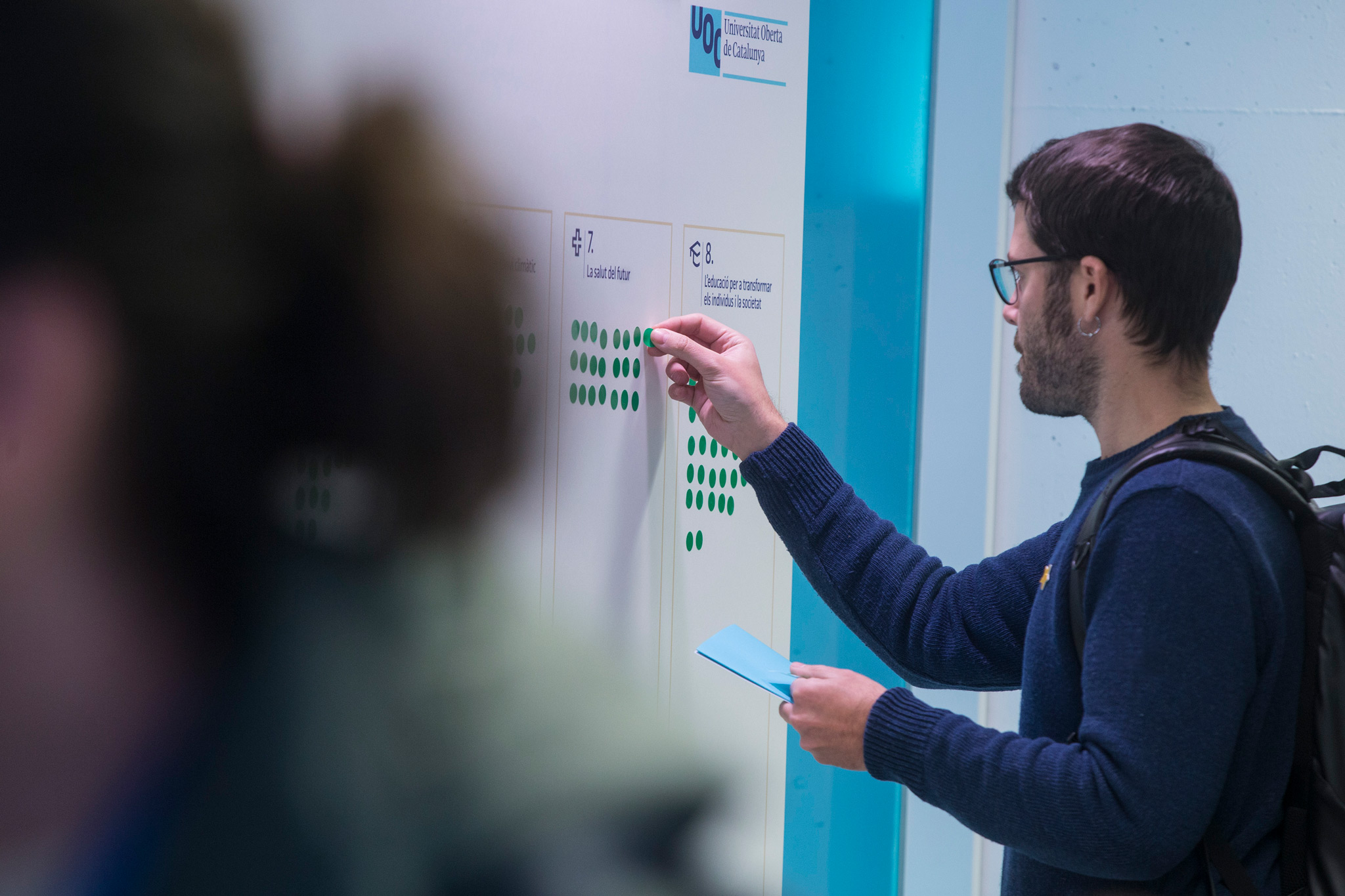 Man wearing glasses placing a green sticker on an interactive panel during a UOC research conference. The words "Universitat Oberta de Catalunya" are visible on the top right of the panel.