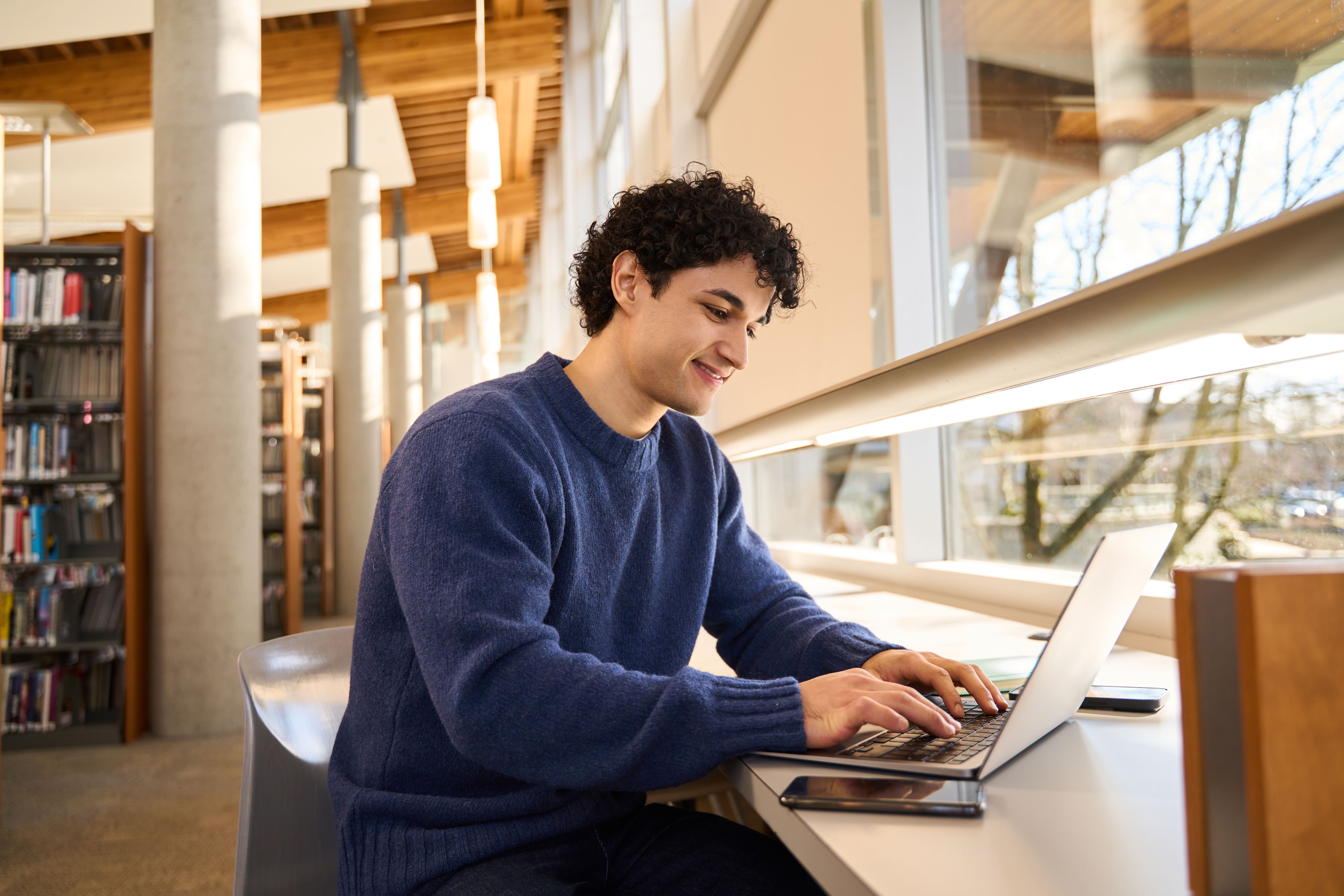 A smiling young student working on a laptop whilst sitting at a desk next to a large window in a brightly lit library.