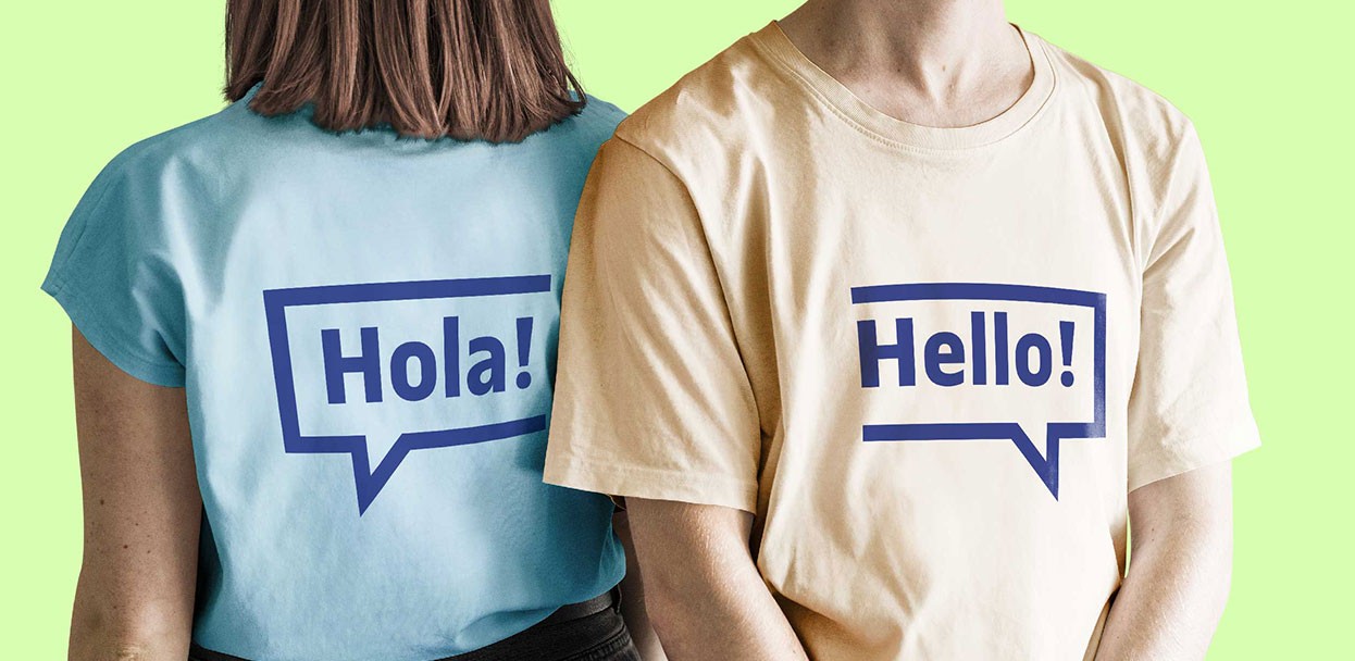 Dues persones d'esquena sobre un fons verd clar, portant samarretes. La de l'esquerra és blava amb una bafarada que diu "Hola!", i la de la dreta és beix amb una bafarada que diu "Hello!".