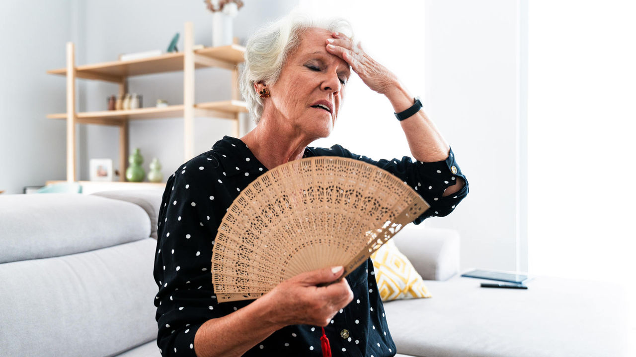 Beautiful senior old woman with gray hair at home using fan to give herself relief against warm climax