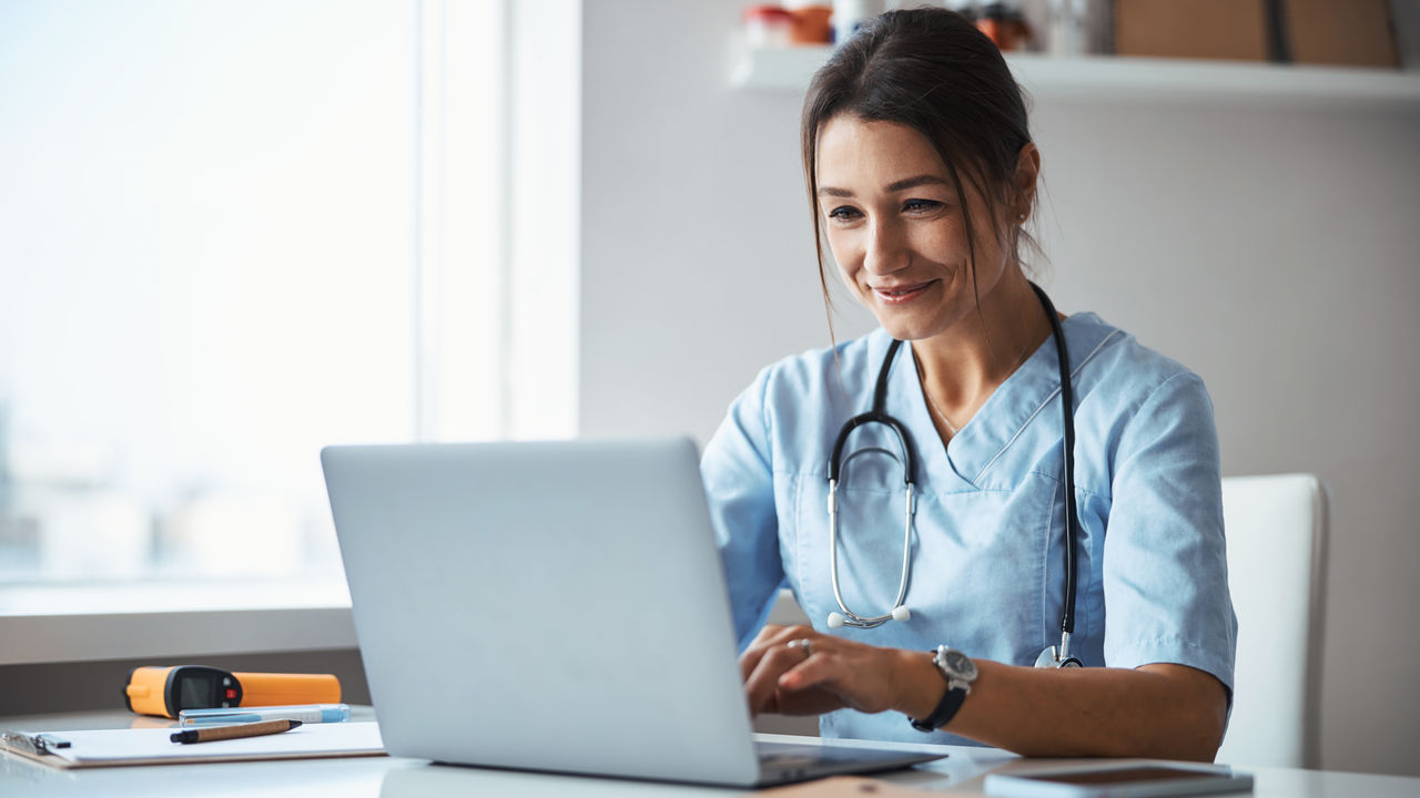 Joyful female doctor using notebook in clinic Beautiful young woman physician sitting at the table and smiling while working on modern laptop
