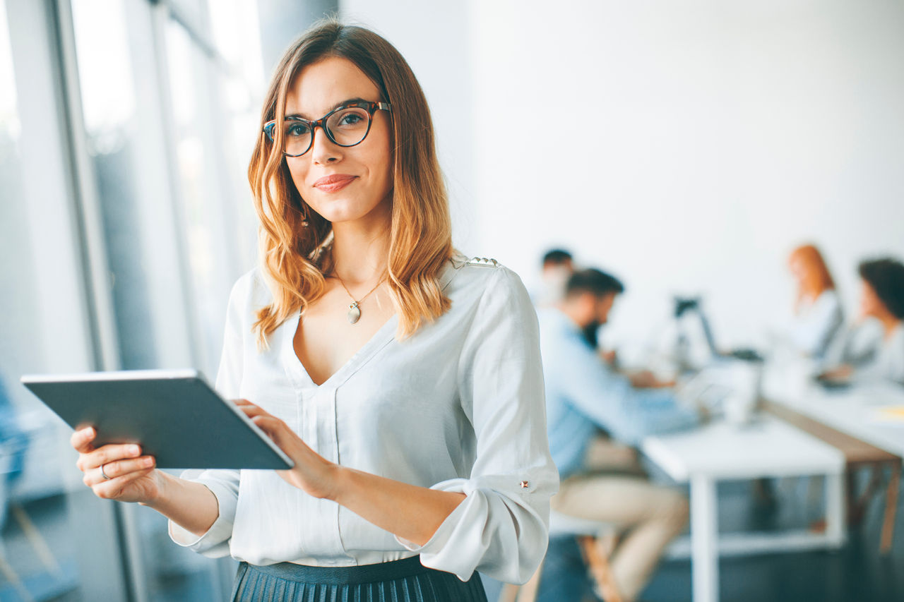 Elegant businesswoman standing in office with digital tablet Young attractive female manager working on digital tablet while standing in modern office